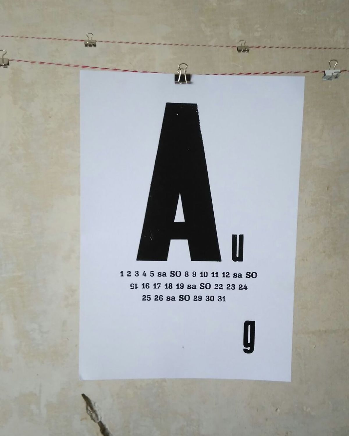 Kalenderblatt August mit großem „A“ und gesetzten Tageszahlen, an einer Leine aufgehängt