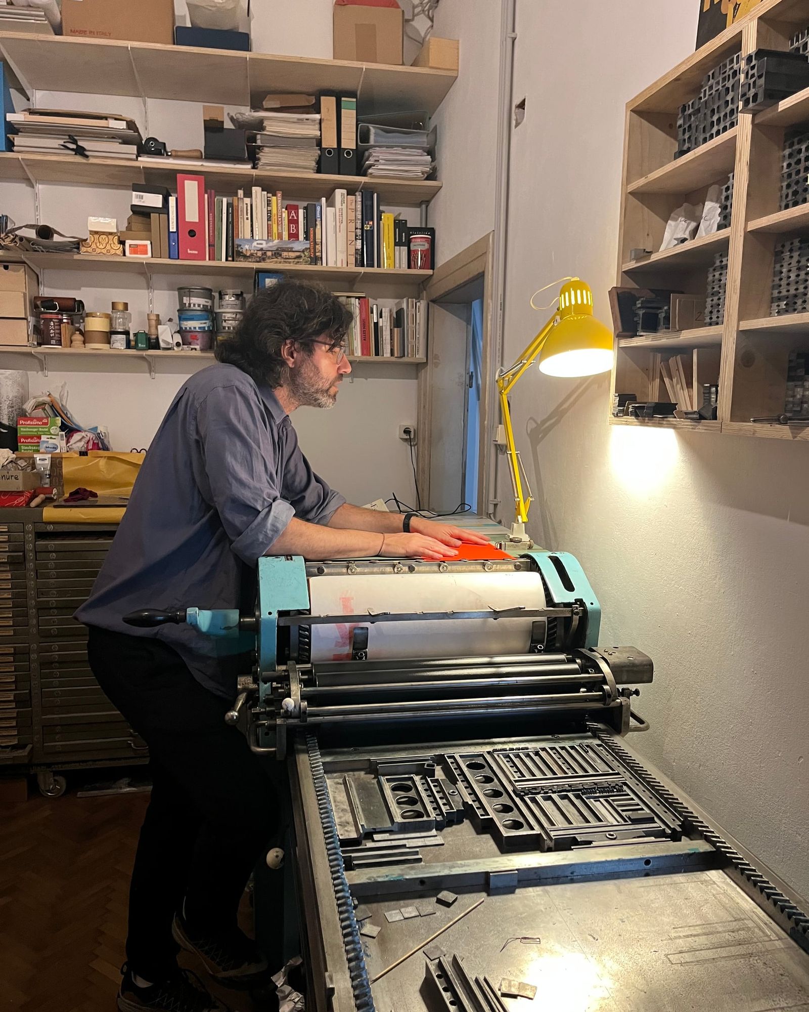 Alexander Zöhrer druckt an der Presse in der Werkstatt, im Hintergrund Bücherregale und eine gelbe Lampe