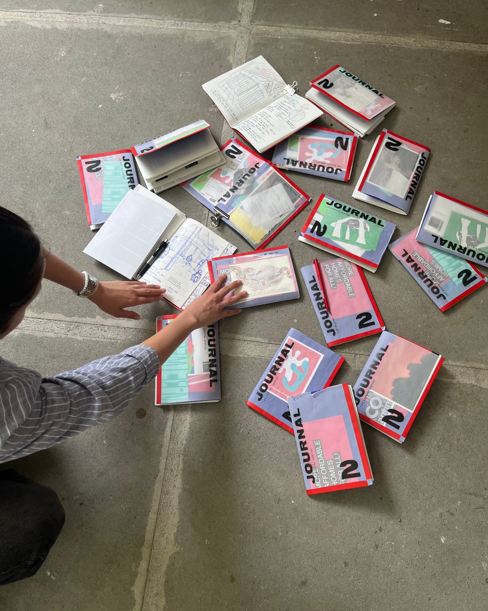 Viele gedruckte Hefte „Journal 2“ liegen ausgebreitet auf dem Boden, eine Hand ordnet sie
